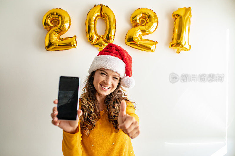 Portrait of a young smiling woman in front of a white wall with a Santa hat holding mobile phone